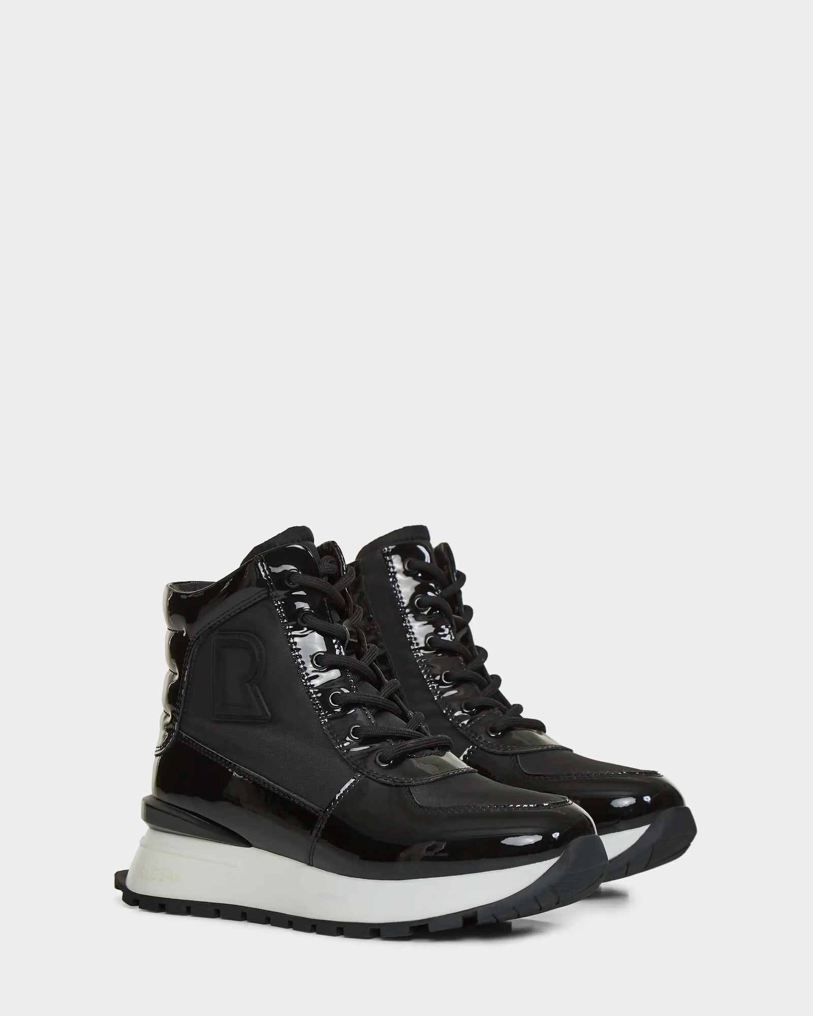 The CHELSA - L223505 Black by Rudsak are black high-top sneakers with chunky white soles, black laces, mixed glossy and matte textures, and a large embossed R on the side—combining bold bottes baskets femmes style with signature RUDSAK design.