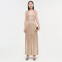 Beige pleated sleeveless gown with deep V-neckline and tiered waistband sections, worn with gold clutch and black sandals.