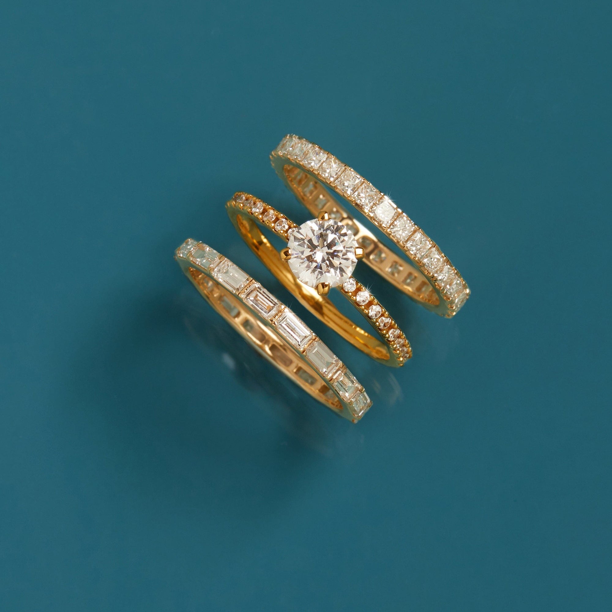 Flatlay Model A stack of multiple gold and diamond rings, including an engagement ring and eternity bands, resting on a dark teal background