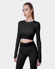 A woman with straight brown hair in a ponytail models the Rudsak L523016T DOREEN - BLACK fitted womens long sleeve sports top and high-waisted black leggings against a plain light grey background.