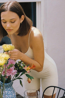 Woman wearing cream ribbed sleeveless midi onesie, touching flowers in a vase.