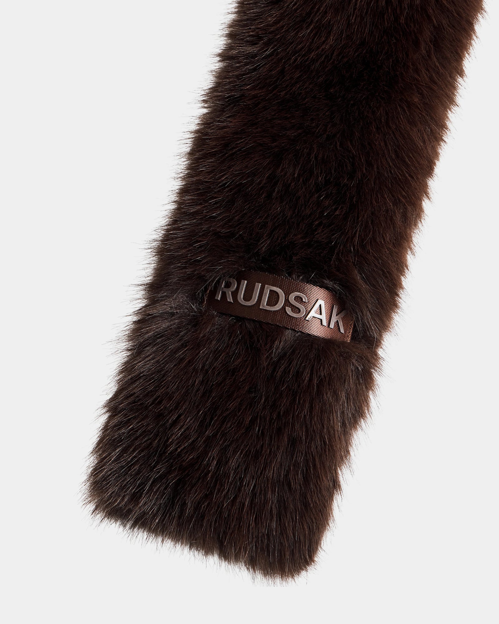 A close-up of the CARMY - L425534 Espresso accessory showcases its dark brown plush texture and warmth, accented by a black leather tag with Rudsak embossed in metallic letters, all set against a plain white background.