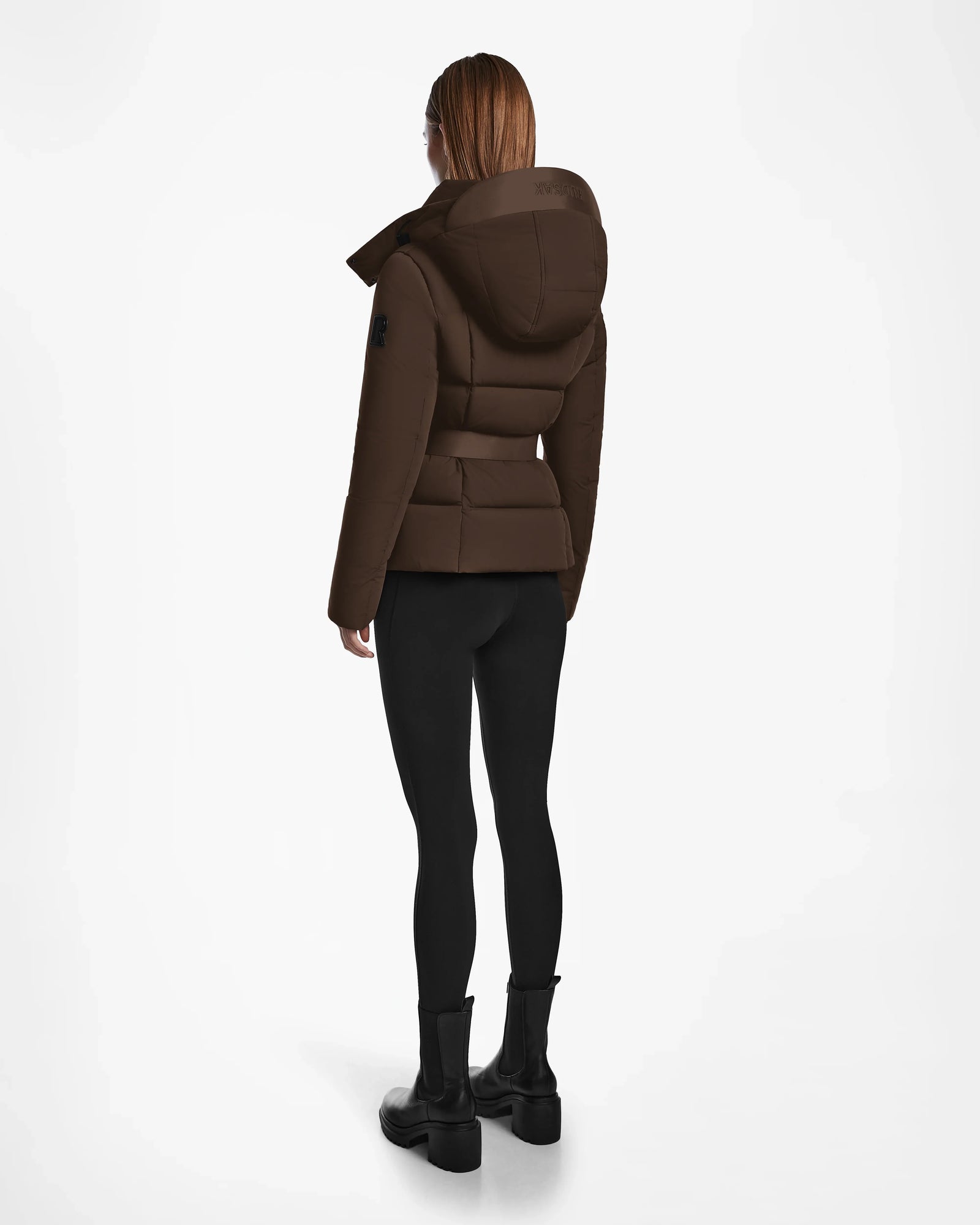 A person with straight, shoulder-length hair stands facing away, wearing the Rudsak ADELINE - 8124544 Mocha quilted short down puffer, black leggings, and black ankle boots against a plain white background.