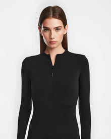 A woman with straight brown hair wears the AGNES - 8524551 Black bodycon stretch dress by Rudsak, featuring a short front zipper. She stands against a plain white background, looking directly at the camera with a neutral expression.