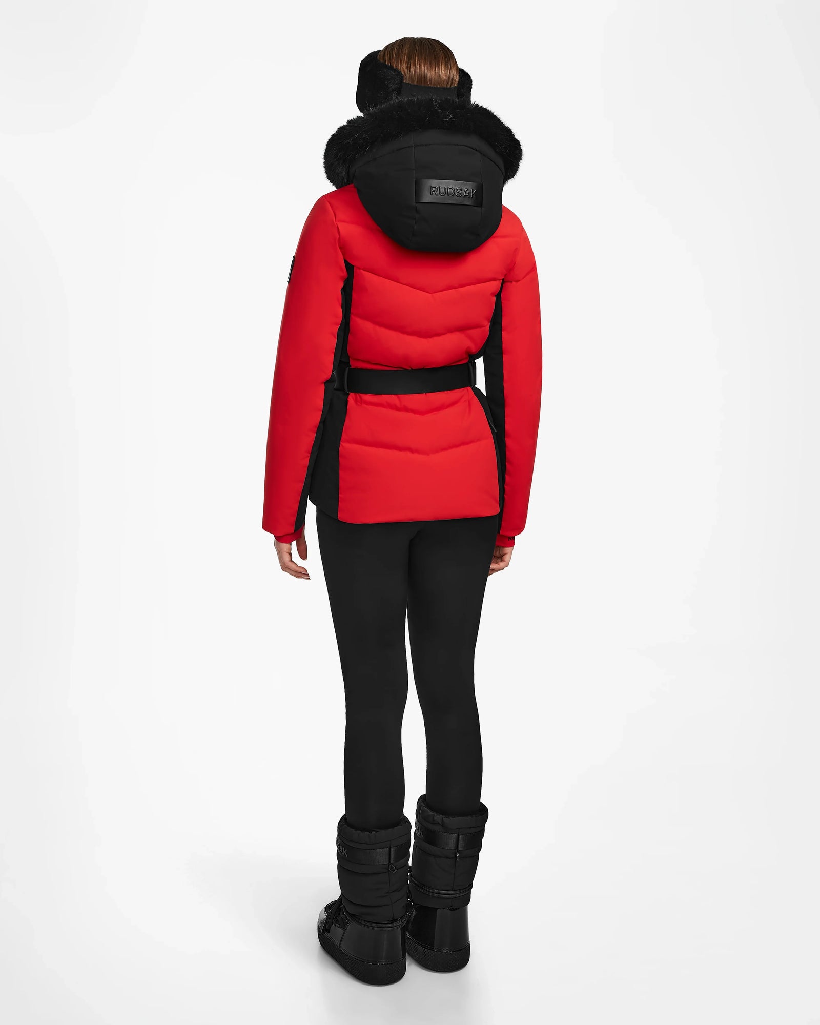 A person stands facing away, wearing the Rudsak ALAIA - 8124816 Flame Red ski jacket with a black belt, paired with black leggings and winter boots against a plain white background.