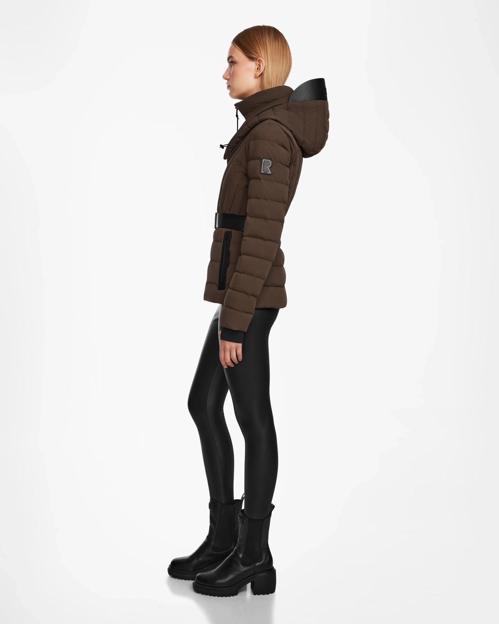 A woman with straight, light brown hair stands in profile wearing the Rudsak ALISA - L124506 Espresso women’s puffer jacket, shiny black leggings, and black ankle boots against a plain white background.