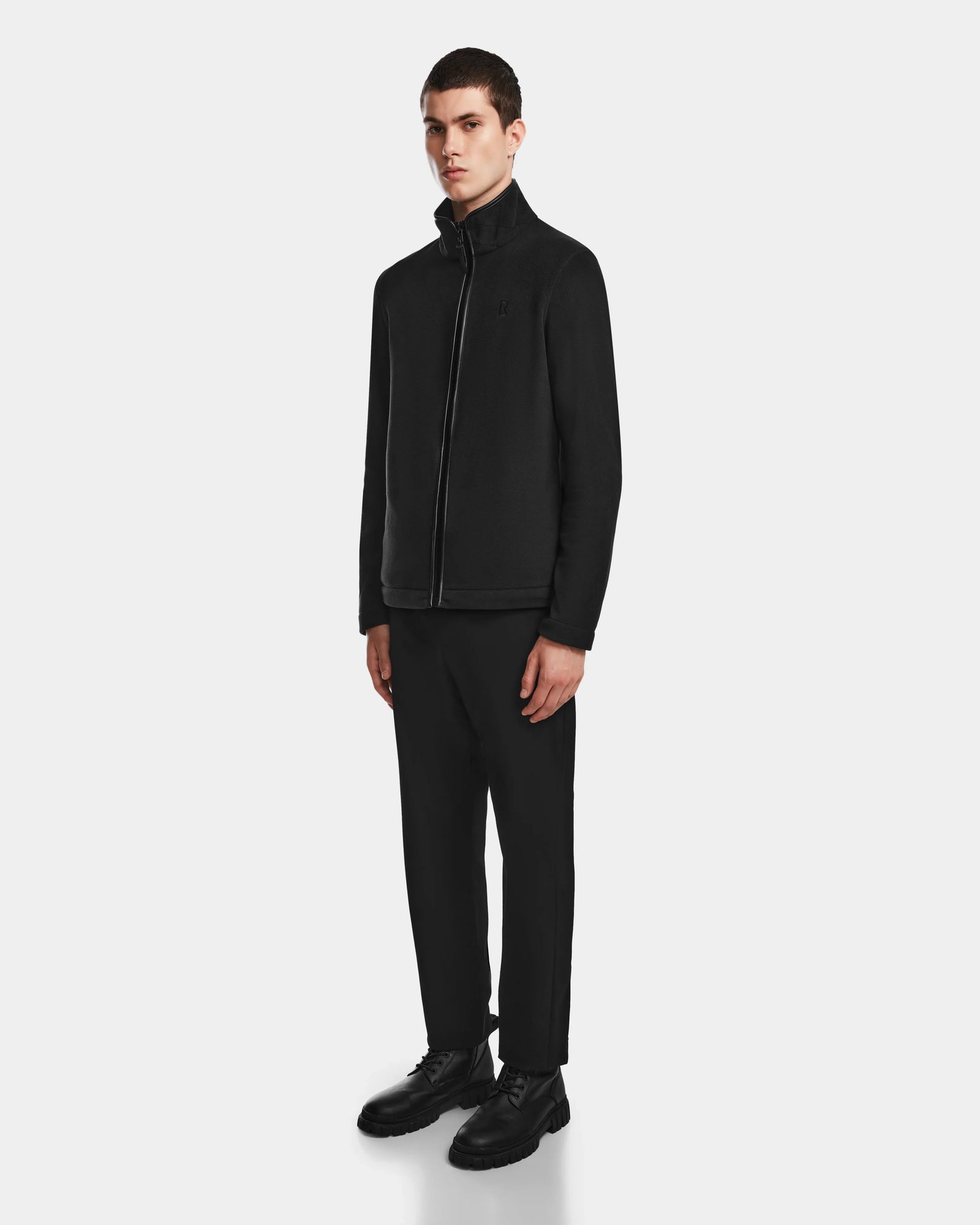 A young man stands against a plain background, wearing the Rudsak ARAGON - 6524539 Black men’s fleece jacket with vegan leather trims, paired with black pants and dress shoes. He faces slightly to the side with a neutral expression.