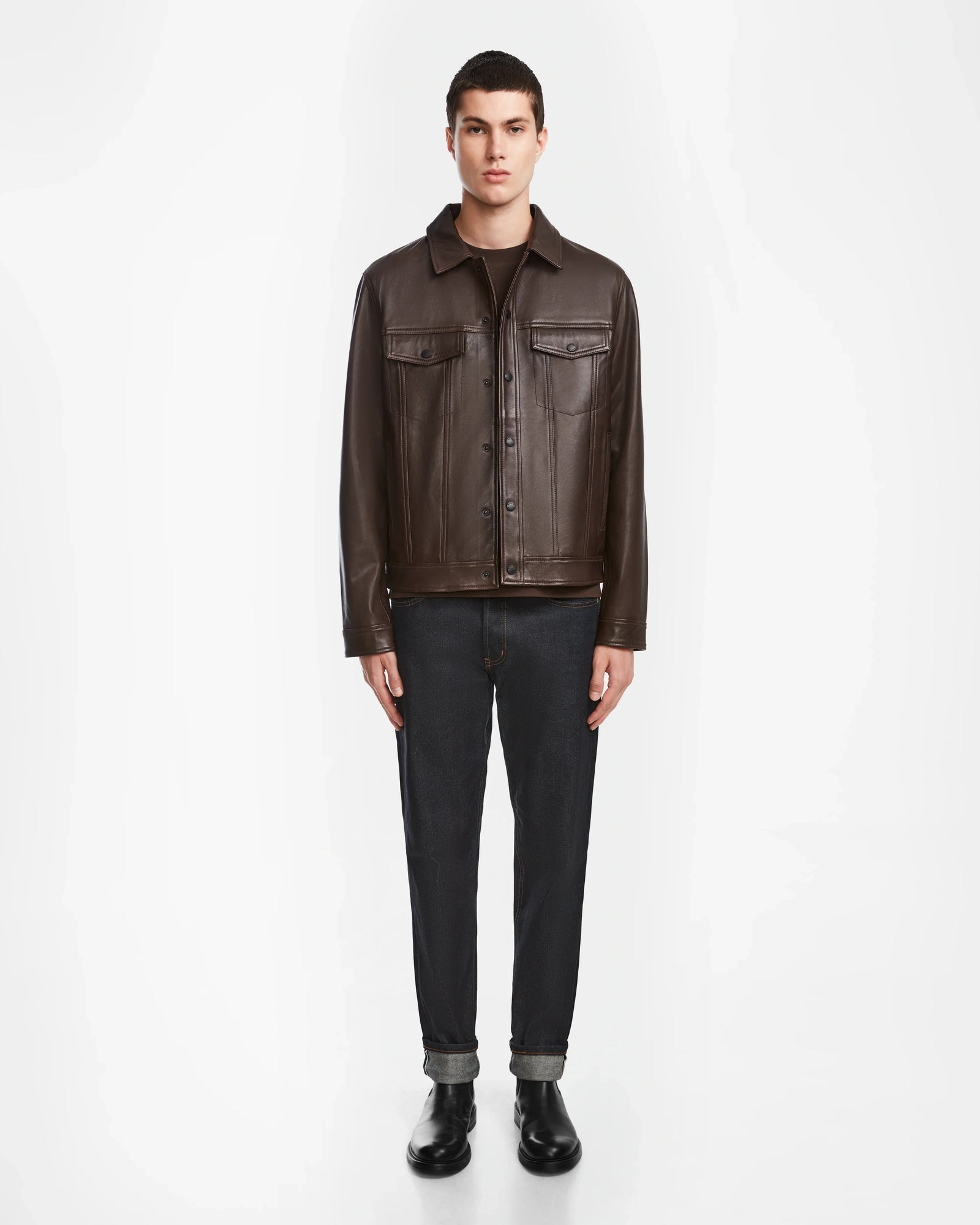 A person stands facing forward against a white background, wearing the Rudsak AREN - 6124580 Espresso mens leather shirt jacket, dark jeans with rolled cuffs, black boots, and a dark shirt.