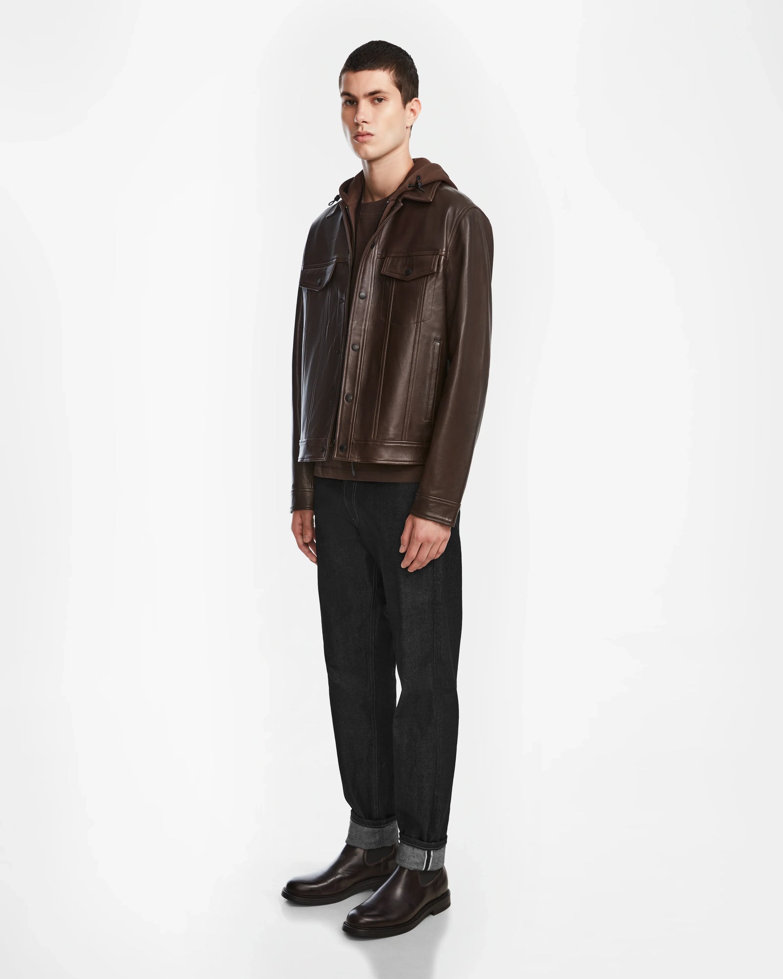 A person stands facing slightly left, wearing the Rudsak AREN - 6124580 Espresso leather jacket over a brown hoodie, black cuffed jeans, and dark brown boots against a plain white background.
