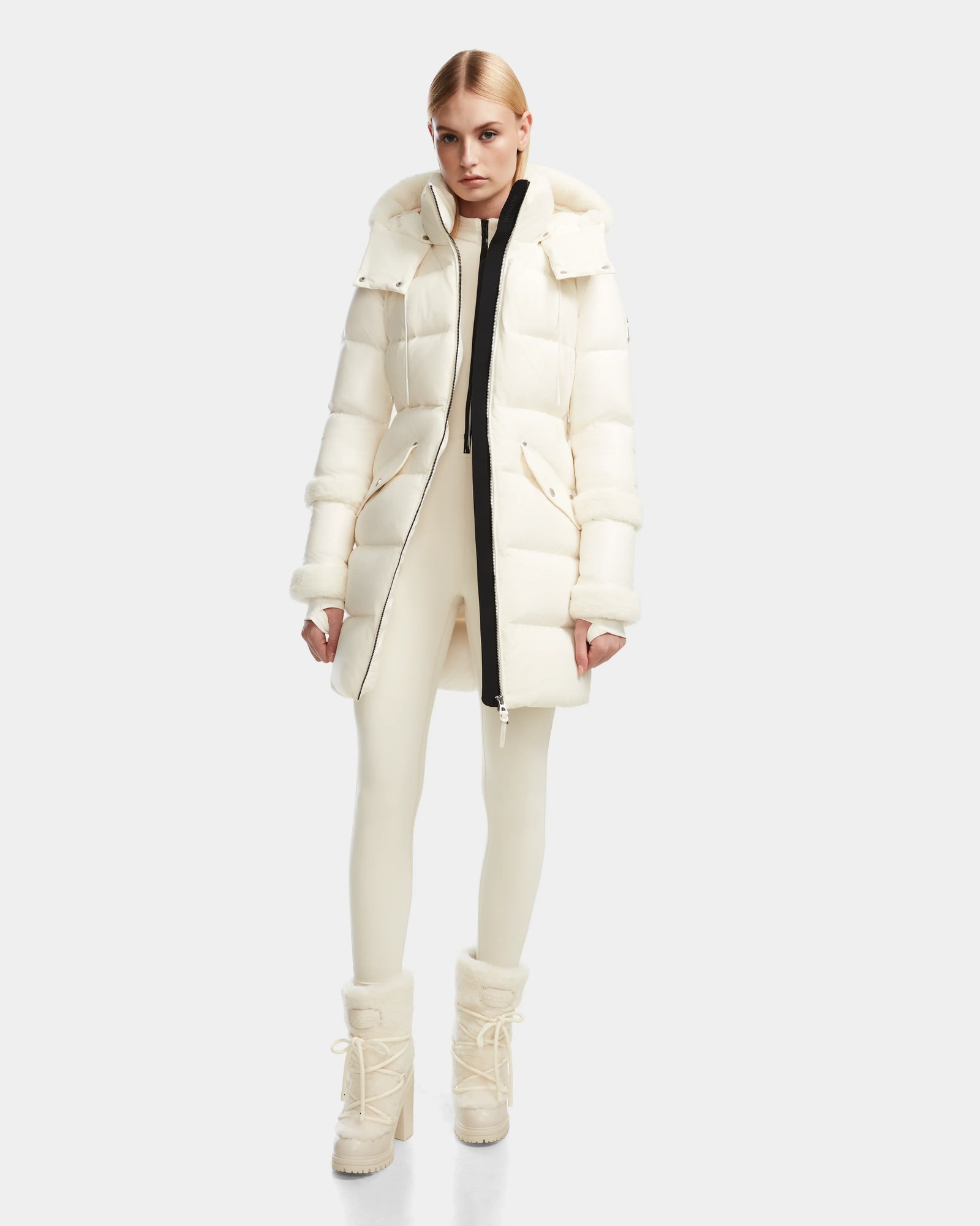 A woman with a serious expression and pulled-back hair models the Rudsak AUDREY L - 8124585 Iceland knee-length down puffer jacket in recycled Stormshell fabric, paired with cream fitted pants and fur-trimmed winter boots.
