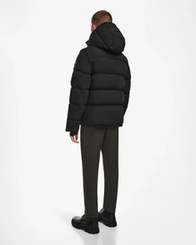 A person stands facing away, wearing the Rudsak BOBBY - 6124506 Black men’s down puffer jacket with a hood, dark pants, and black shoes against a plain white background.