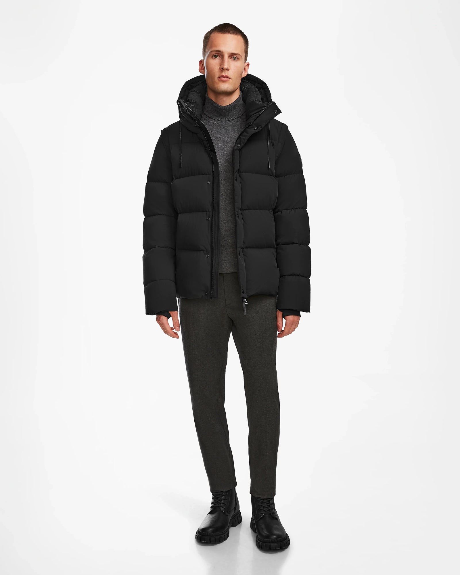A man stands against a white background wearing the Rudsak BOBBY - 6124506 Black mens down puffer, a gray turtleneck, dark pants, and black boots, facing forward with a neutral expression.