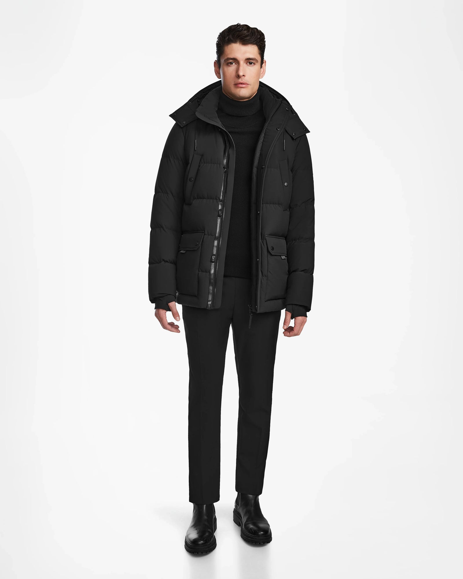 A man wearing the Rudsak CARLOS - M124509 Black parka stands against a white background paired with black turtleneck, pants, and boots, facing forward with a neutral expression and hands relaxed at his sides.