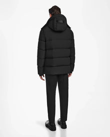 A man stands facing away, wearing the Rudsak CARLOS - M124509 Black men’s down parka with a hood, along with black pants and shoes, against a plain white background.