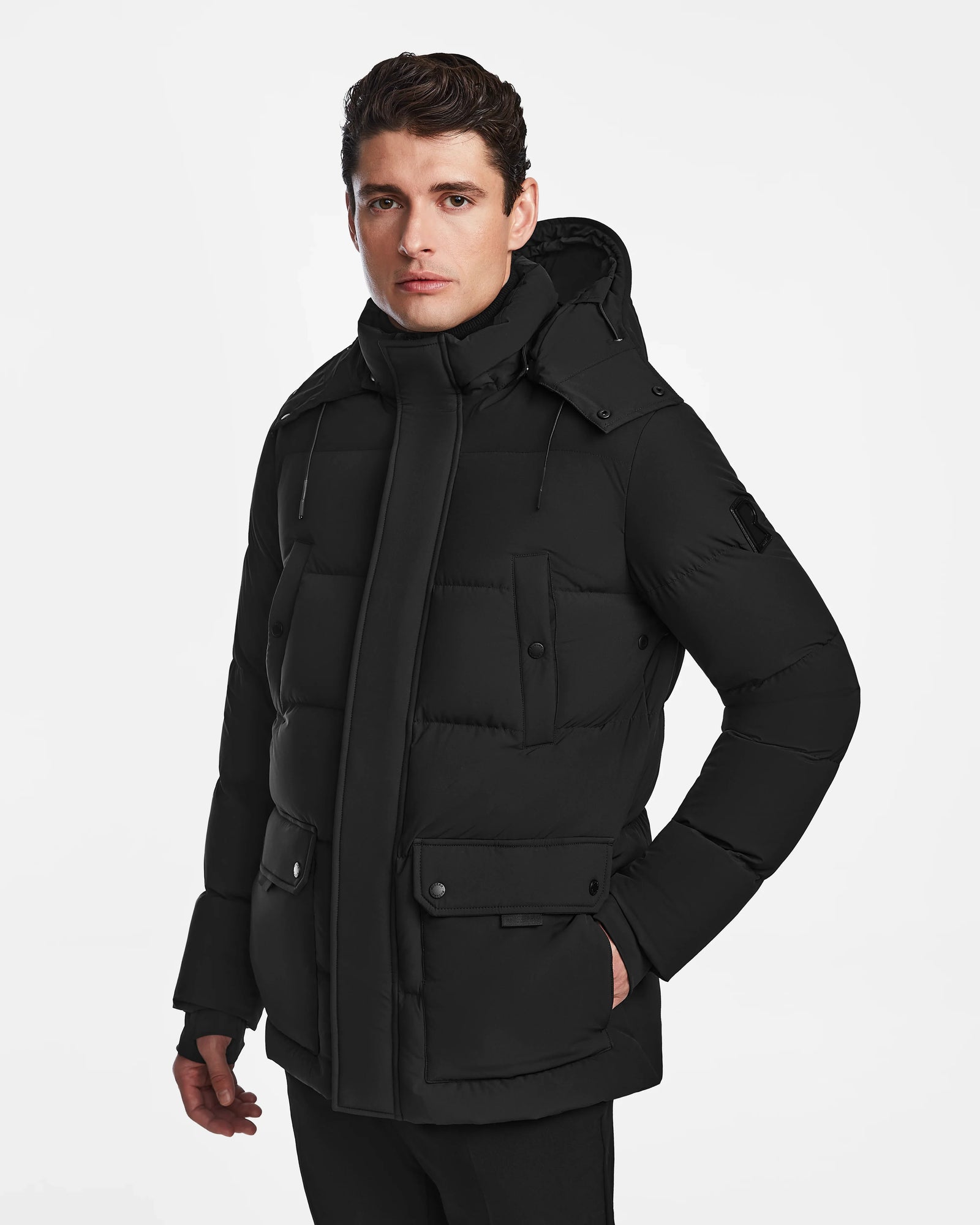 A man with short brown hair wears the Rudsak CARLOS - M124509 Black quilted parka with a hood and front pockets, standing against a plain white background.