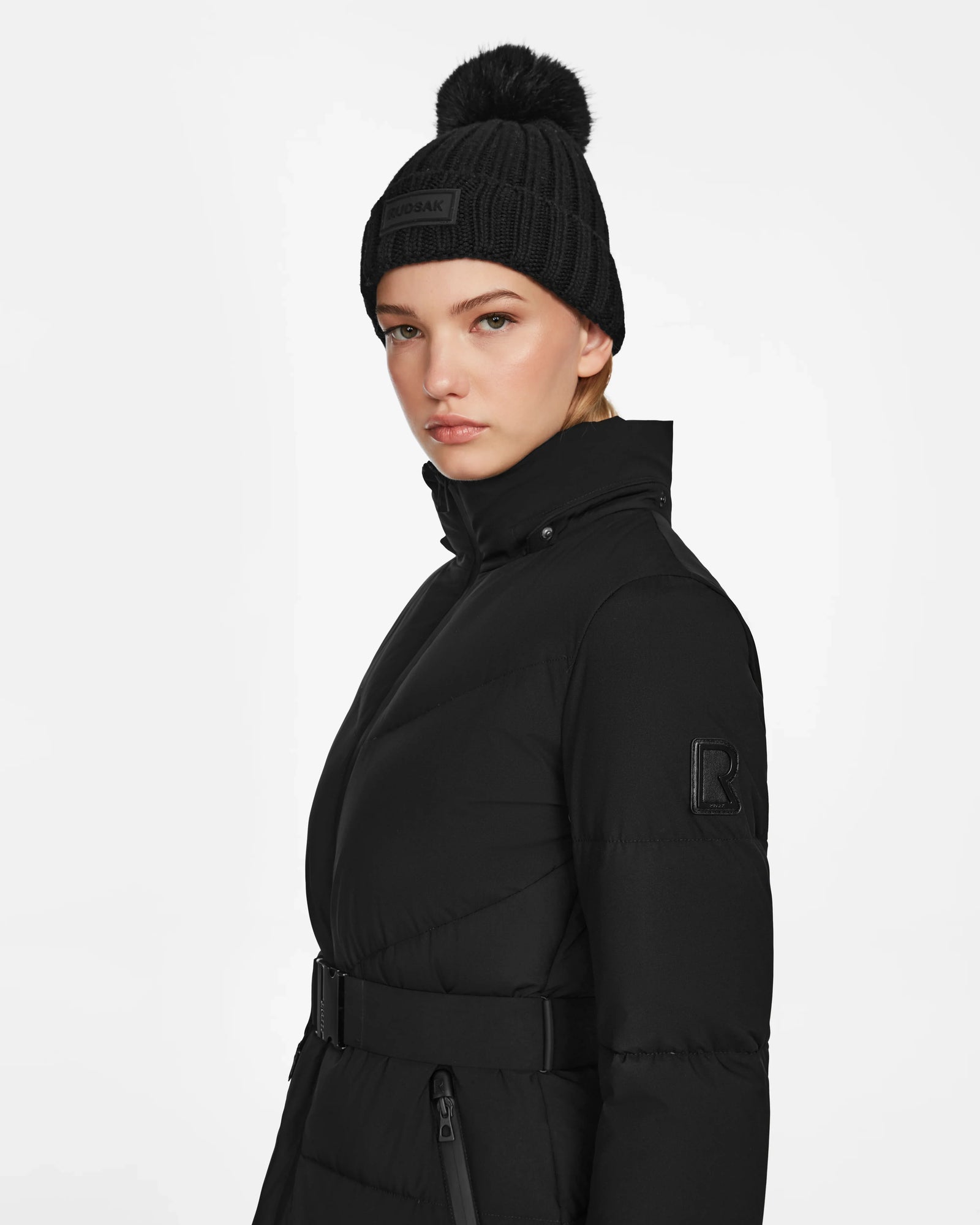 A woman wears the Rudsak CHRISTEL M-M - L124505 Black quilted winter coat and a pom-pom beanie, standing against a plain white background with a neutral expression, looking slightly to the side.