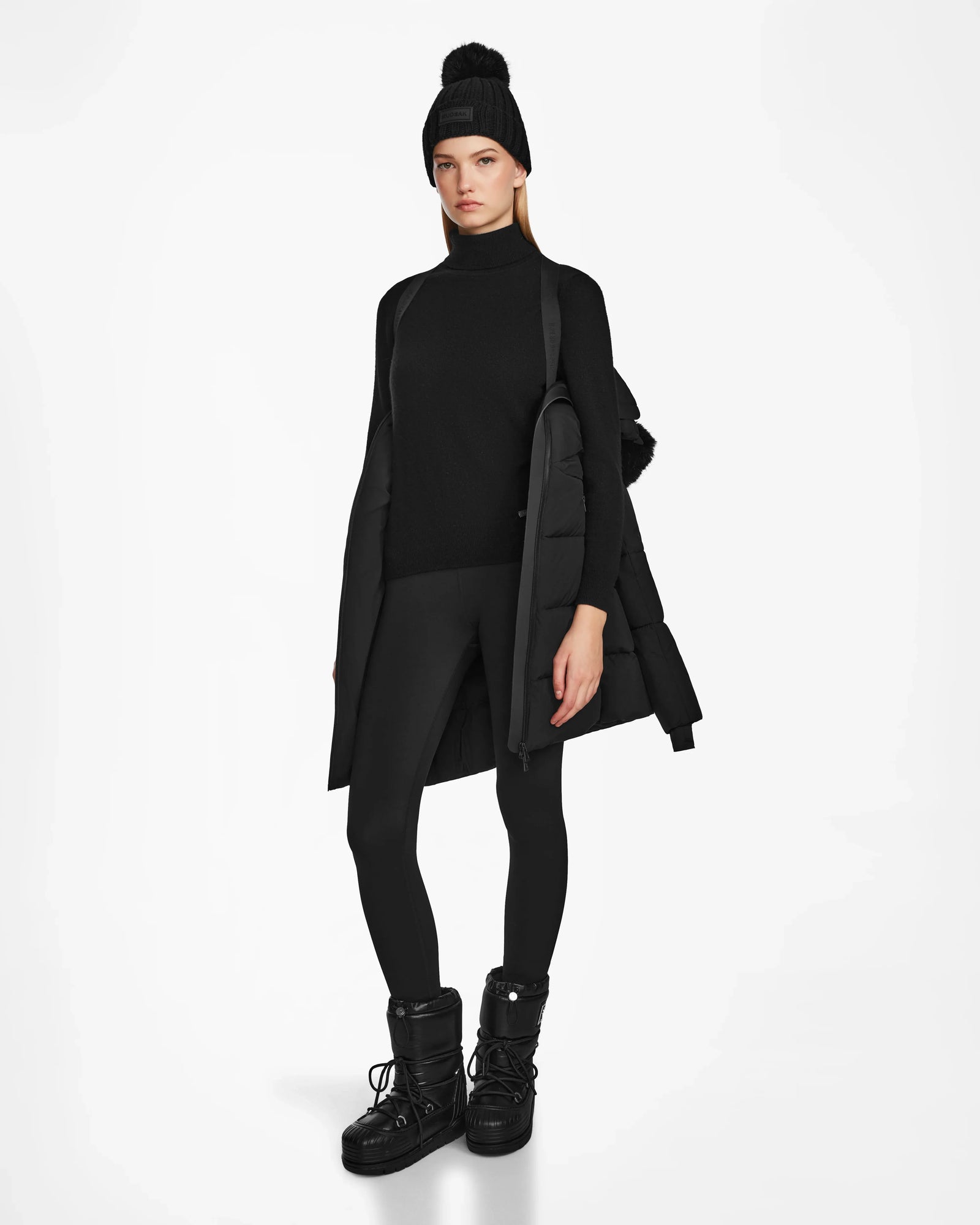 A person with a neutral expression stands against a white background wearing the Rudsak CHRISTEL M-M - L124505 Black quilted winter coat draped over their shoulders. They’re also dressed in a black beanie, turtleneck, leggings, and winter boots.