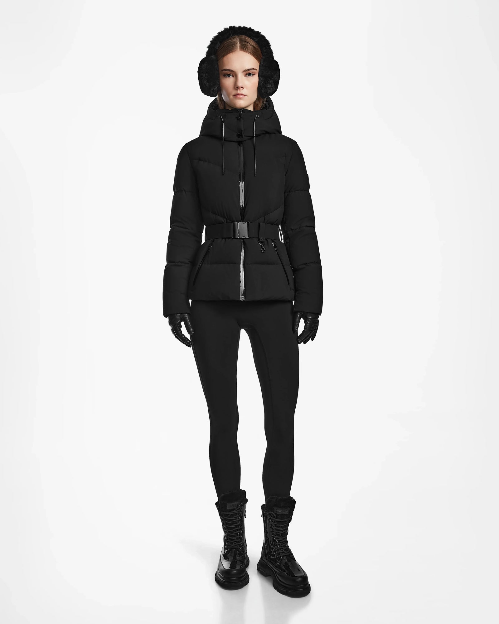 A woman stands against a white background wearing the Rudsak CHRISTELLE M - L123525 Black quilted down puffer jacket with a belt, paired with matching earmuffs, gloves, leggings, and lace-up boots for a fitted look.