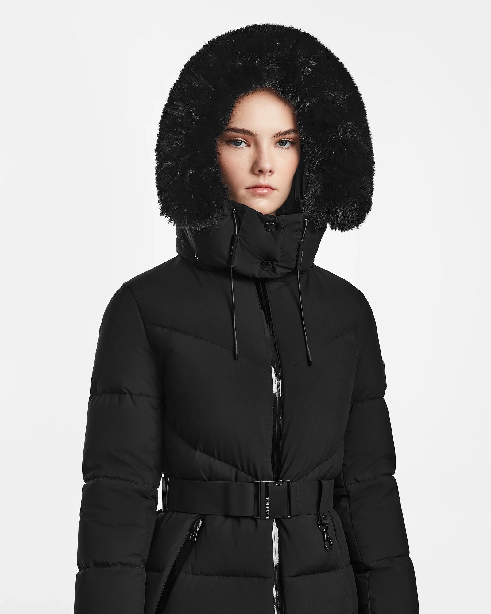 A woman wearing the Rudsak CHRISTELLE M - L123525 Black sustainable winter coat with a fitted silhouette and large fur-lined hood stands against a white background, looking straight ahead with a neutral expression.