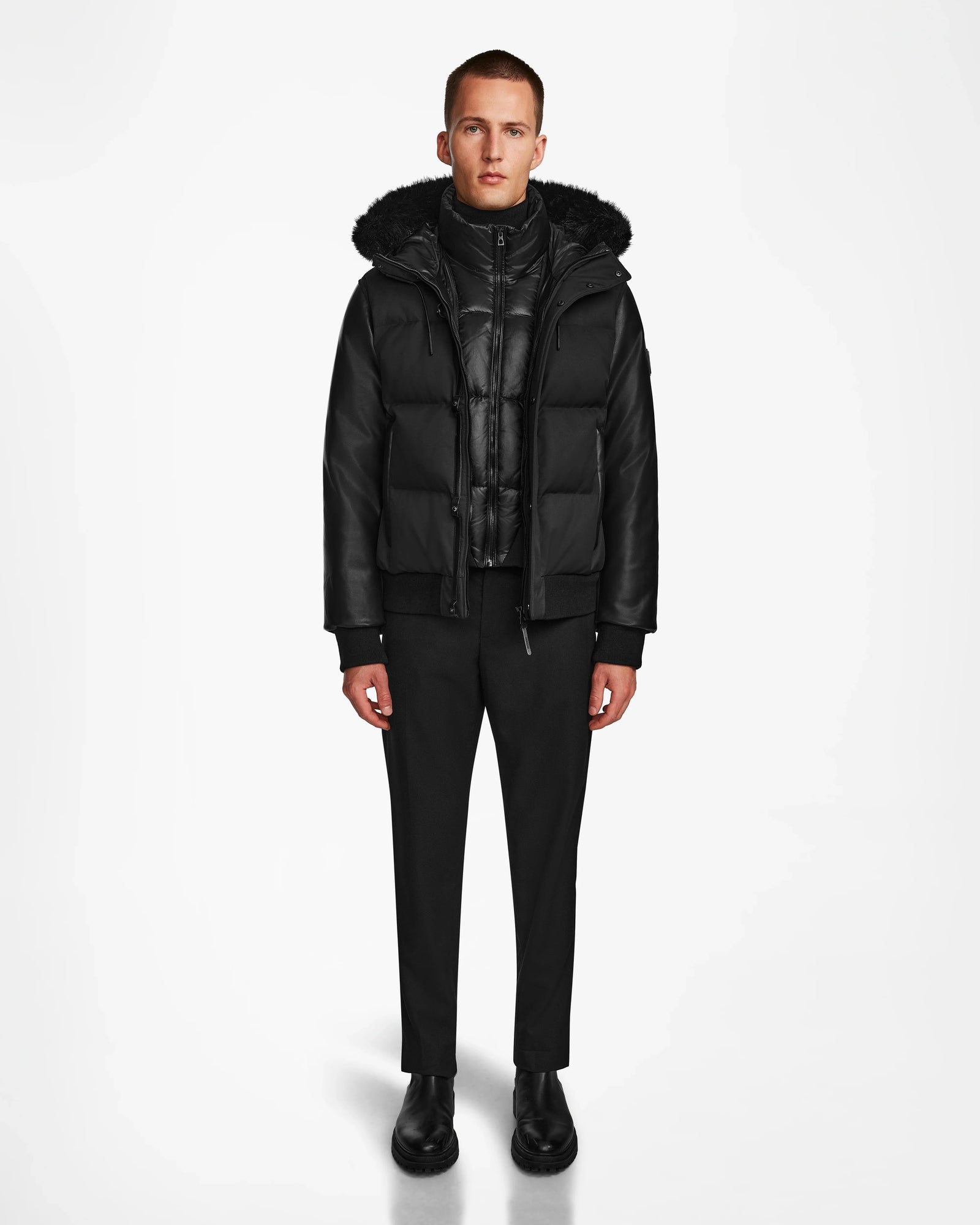 A person stands facing forward against a plain white background, wearing the Rudsak DALLAS R - M123501 BLACK— a weather-resistant hooded puffer jacket with recycled leather sleeves—paired with black pants and shoes.