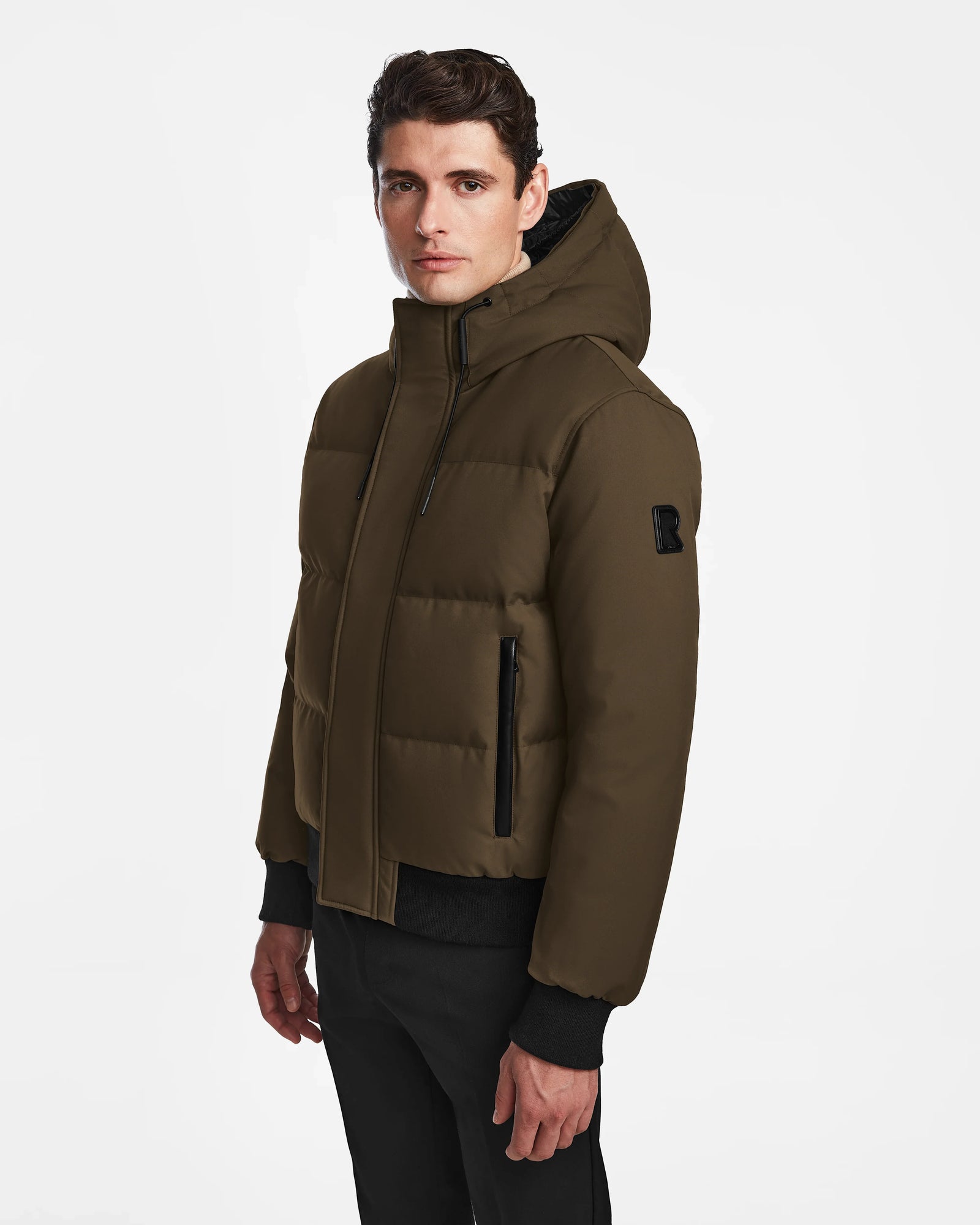 A young man wears the Rudsak DALLAS - M122501 Army, a weather-resistant puffer jacket with black zippers and black pants, standing against a plain white background and looking slightly to the side.