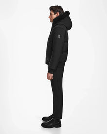 A man in profile wears the Rudsak DALLAS - M122501 Black weather-resistant puffer coat with fur trim, paired with black pants and boots, set against a plain white background.