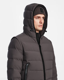 A man wears the Rudsak DEMIAN M-R - M123504 Grey down puffer jacket with a hood up and a ribbed knit beanie, made from sustainable recycled fabric, standing against a plain white background.
