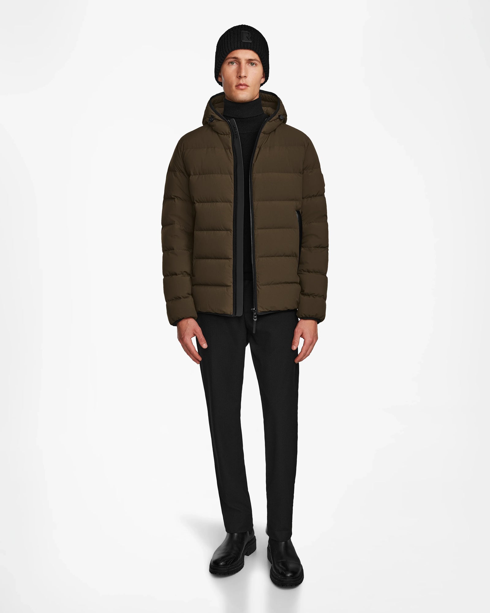 A person stands against a plain white background wearing the Rudsak DEMIAN M - M123507 Army men’s down puffer jacket, made from sustainable recycled fabric, paired with black pants, boots, sweater, and a knit beanie.