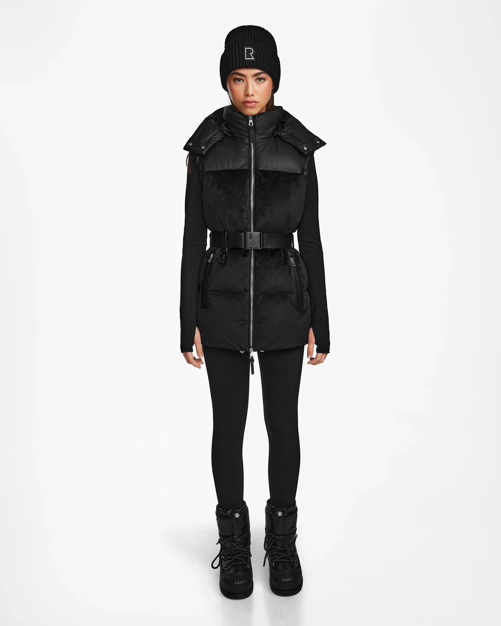 A woman faces forward in an all-black winter look featuring the Rudsak DONIA - L124563 Black down puffer vest, long-sleeve top, leggings, beanie, and winter boots against a plain white background.