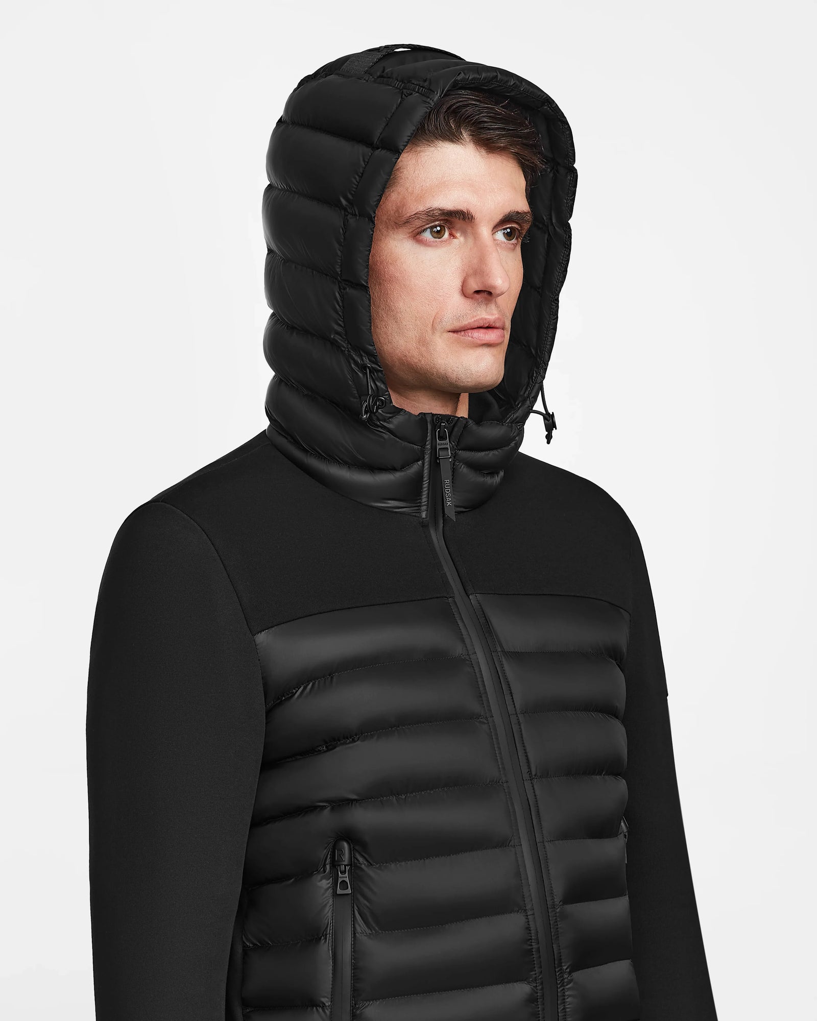 A man wearing the Rudsak DURIN M - M123515 Black mens hooded jacket with lightweight down insulation, zipper, and side pockets looks slightly to the left against a plain white background.