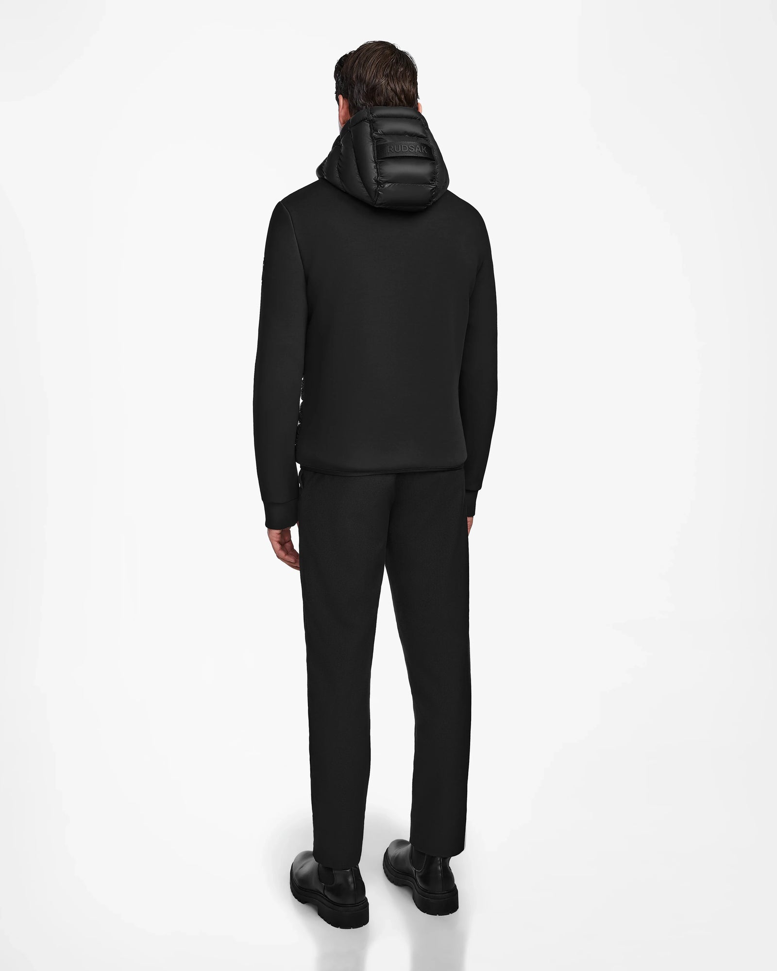 A man stands facing away, wearing Rudsaks DURIN M - M123515 Black mens hooded jacket with lightweight down insulation, paired with dark pants and black shoes, set against a plain white background.