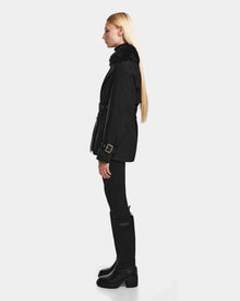 A woman with long blonde hair stands in profile wearing the Rudsak ELENOR - 8124707 Black womens trench coat, styled with black pants and knee-high boots, against a plain white background.