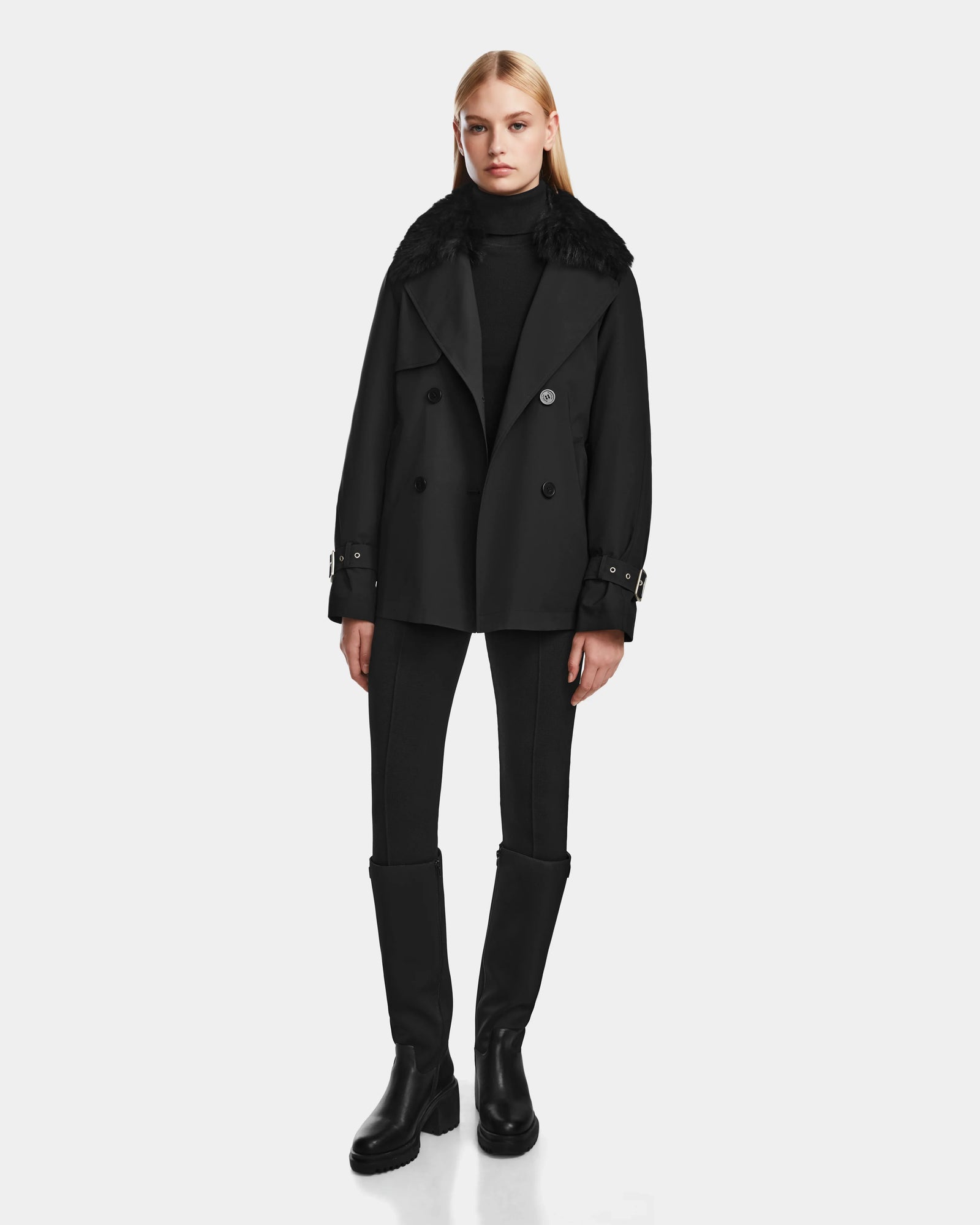 A woman faces forward against a white background, wearing the Rudsak ELENOR - 8124707 Black double-breasted women’s trench coat with a wide shearling collar, styled with a black turtleneck, fitted pants, and knee-high boots.