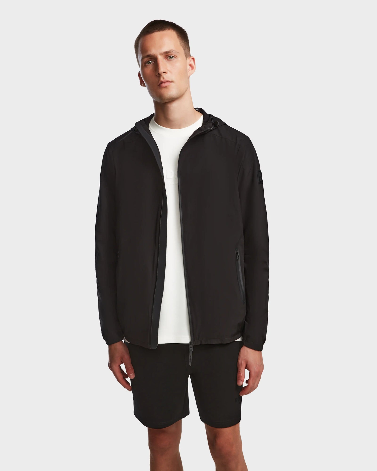 A man stands against a plain background wearing the Rudsak ELIJAH - M124000 Black hooded zip-up jacket over a white t-shirt and black shorts, with his arms relaxed by his sides.