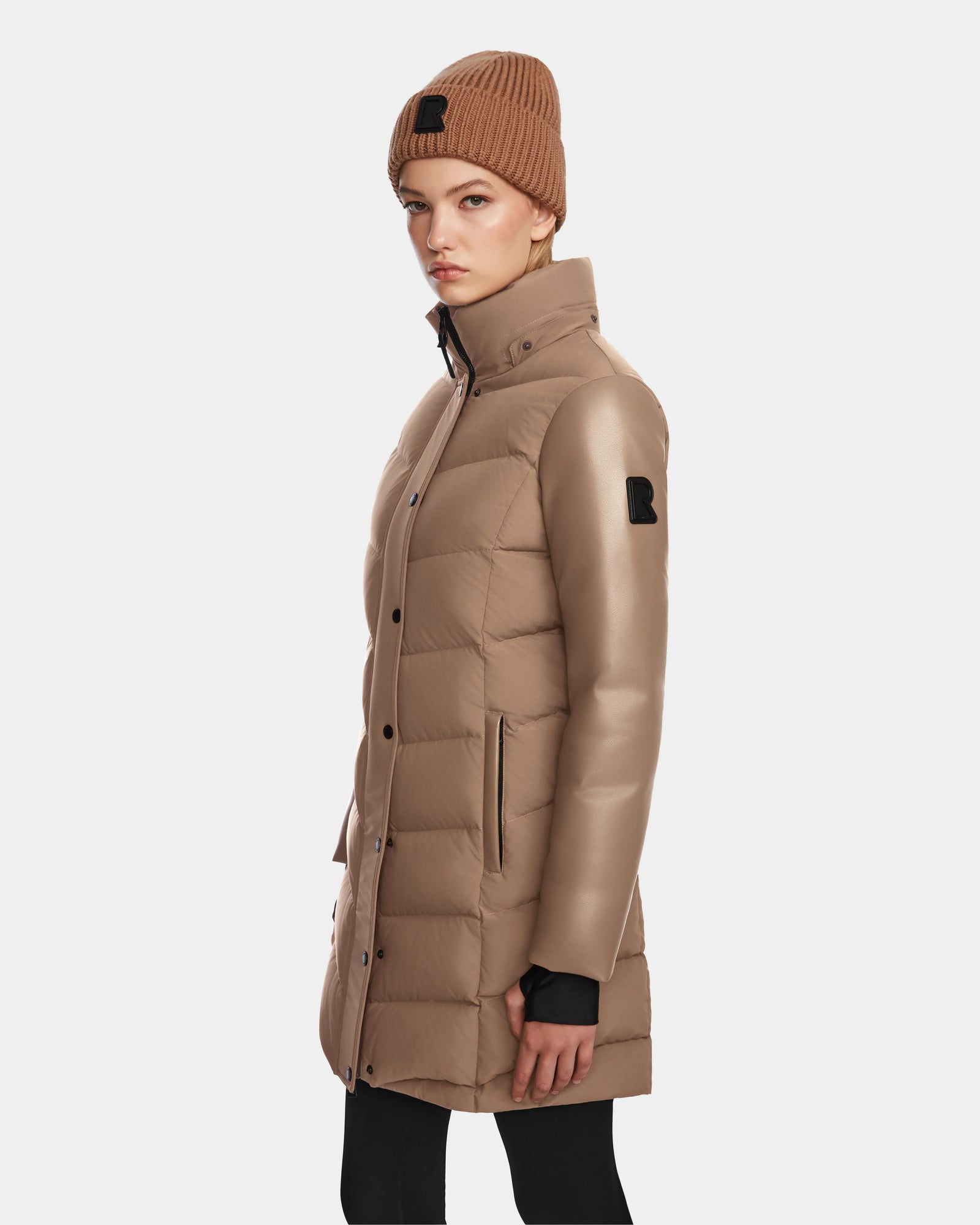 A woman wearing the Rudsak ELSA - L123535 Sand women's quilted down puffer coat and a matching brown knit beanie stands against a plain white background, looking at the camera with a neutral expression.