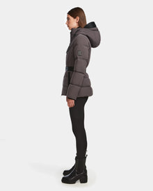 A woman stands in profile wearing the Rudsak ENZAH - 8124692 Grey RDS-certified down women’s puffer jacket with a black belt, black leggings, and ankle boots against a plain white background.