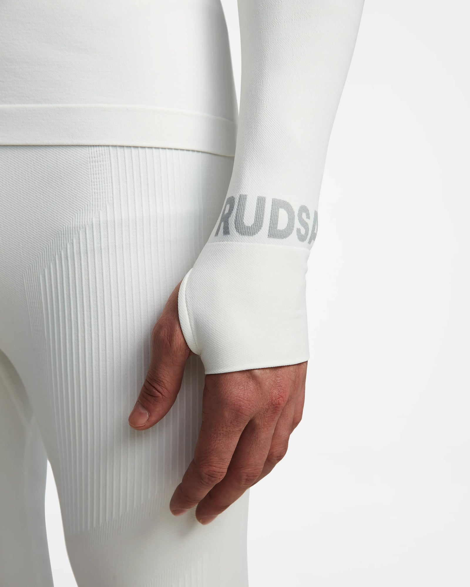 A person wears Rudsaks FRANC - 6524541 Iceland white long-sleeve stretch base layer and matching textured leggings, with a hand through the sleeve’s thumb hole and faint gray text near the wrist visible.