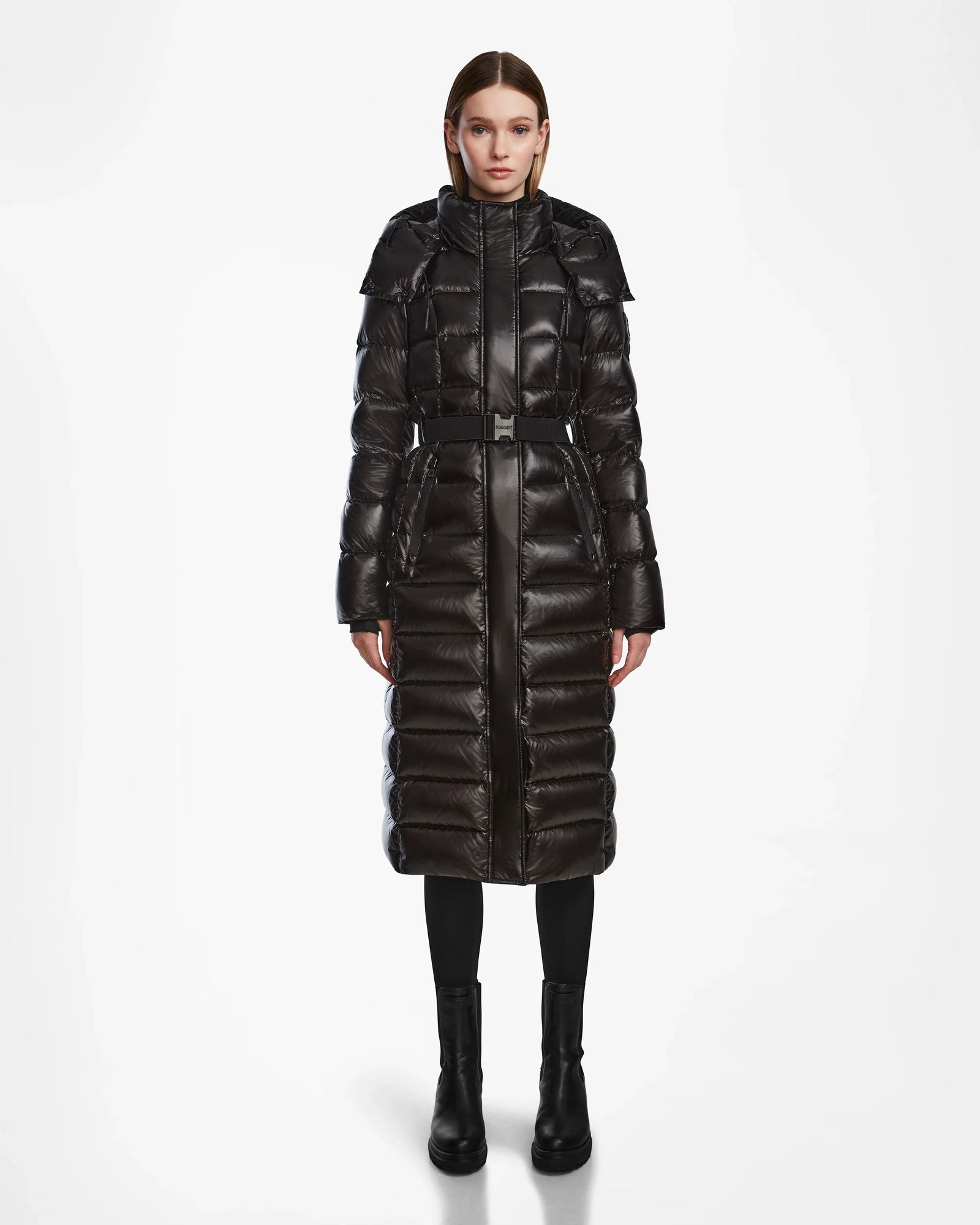 A woman faces forward, modeling the Rudsak FRIDA L-S - L124525 Black, a women's quilted maxi down puffer with high collar and belt, paired with black boots. The plain white background highlights this winter essential.