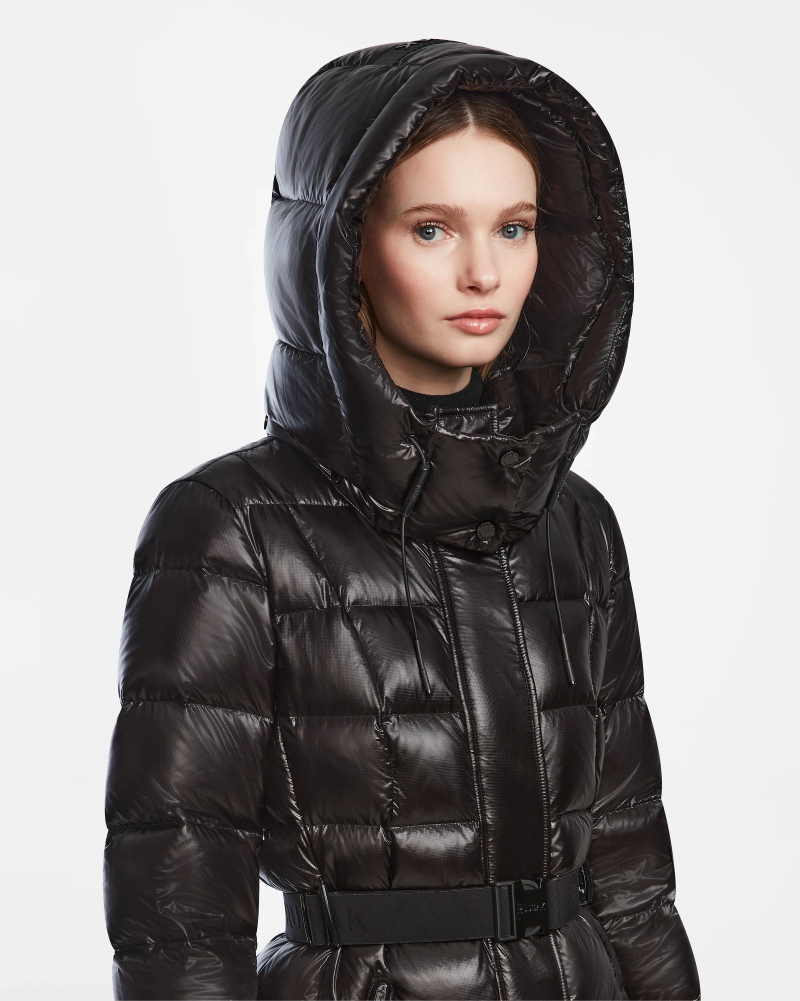 A woman models the Rudsak FRIDA L-S - L124525 Black belted quilted puffer with a large hood against a white background. The shiny black coat adds chic style to any winter wardrobe as she gazes to the side with a neutral expression.