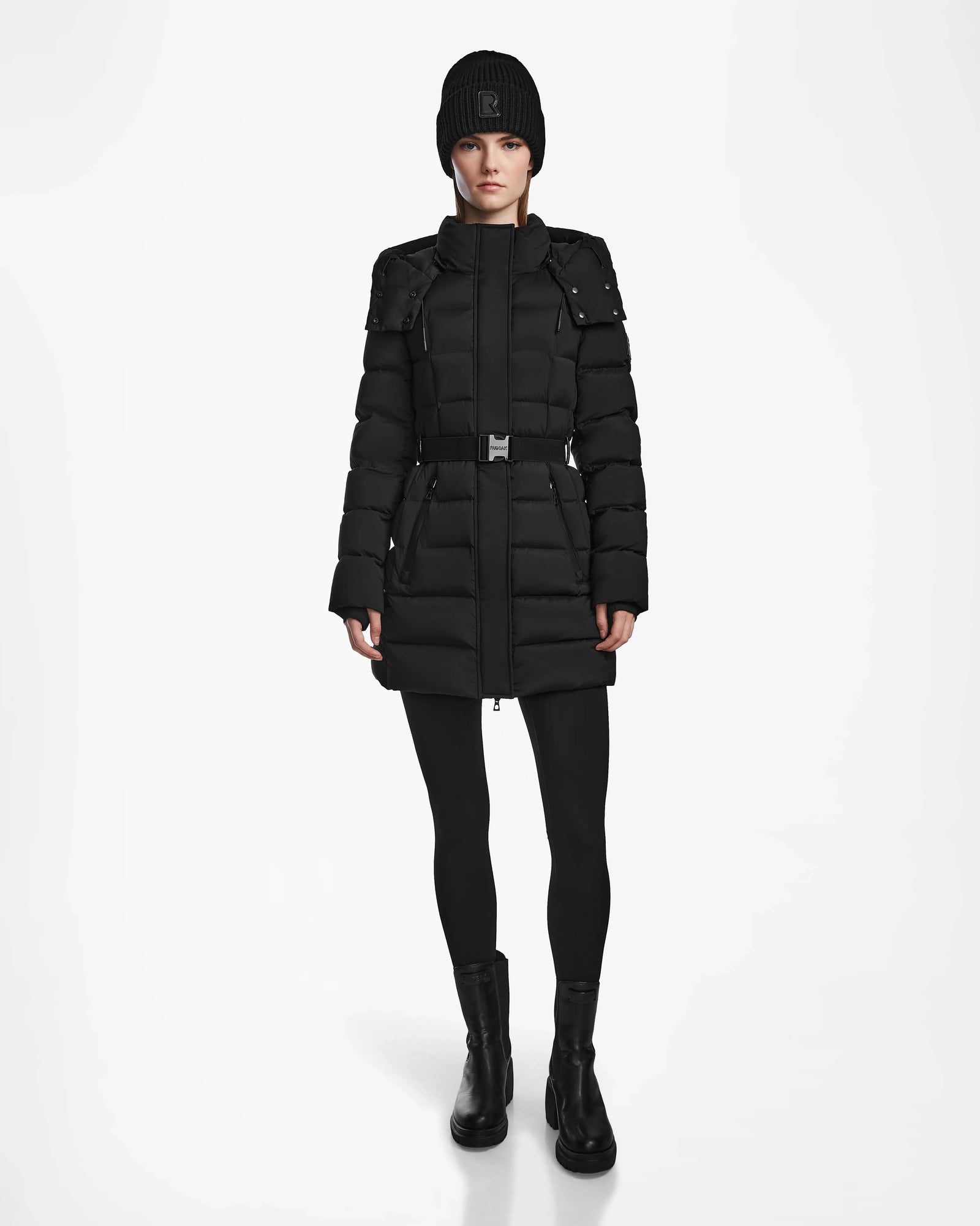 A person wearing the Rudsak FRIDA M - L124524 Black women’s mid-length down jacket with a belt, black tights, boots, and a knit beanie stands facing forward against a plain white background.