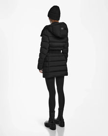 A person wears the Rudsak FRIDA M - L124524 Black womens mid-length down jacket, paired with black leggings, boots, and a knit beanie, standing facing away against a plain white background.