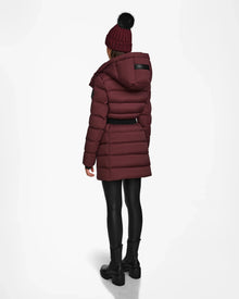 A person wearing the Rudsak FRIDA M - L124524 Wine mid-length down jacket with a removable hood, black leggings, black boots, and a maroon knit hat with a black pom-pom stands with their back to the camera on a white background.