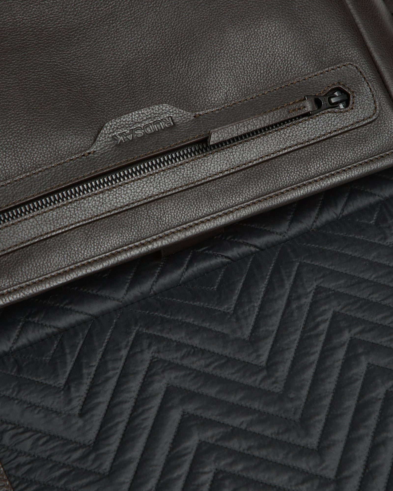 Close-up of the Rudsak GRAYSON - 8324512 Espresso brown leather crossbody bag with a zipper pocket and adjustable strap, shown atop black quilted fabric with a zigzag pattern.