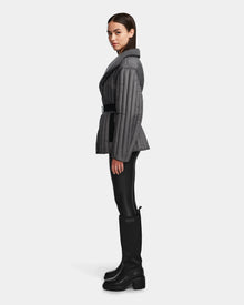 A woman in profile wears the Rudsak INES T - 8124625 Grey quilted jacket with a belt, paired with black leggings and tall black boots, set against a plain white background.