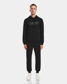 A man faces forward against a white background, wearing the black Rudsak JARED - 6524533 hoodie and matching jogger pants, paired with white sneakers.