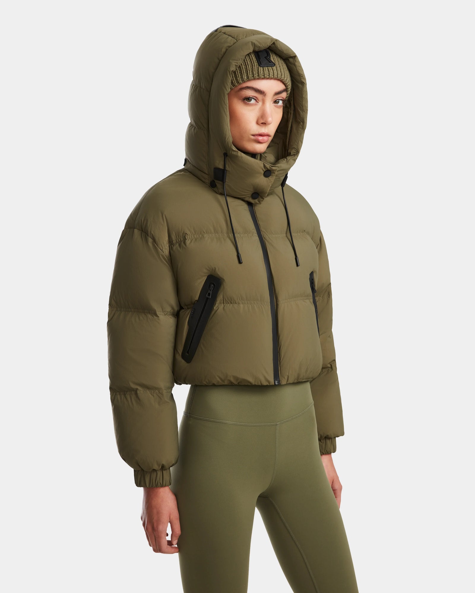 A woman wears the Rudsak KARLEE - 8124721 puffer in Loden Green, styled with matching leggings and a knit beanie, standing against a plain white background.