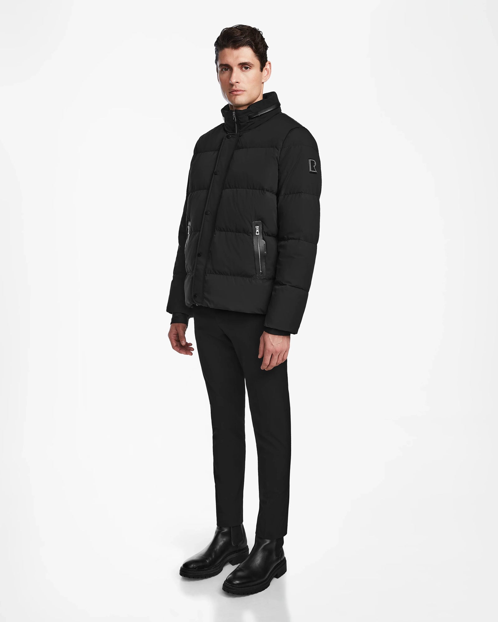 A man stands against a plain white background wearing the Rudsak KARLS - M124505 Black men’s puffer jacket, paired with black pants and boots. He faces forward with a neutral expression, arms relaxed at his sides.