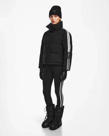 A person stands against a plain white background wearing the Rudsak KIMIA - 8124716 Black women's ski jacket with white sleeve stripes, paired with a black beanie, black gloves, black leggings with white side stripes, and black winter boots.