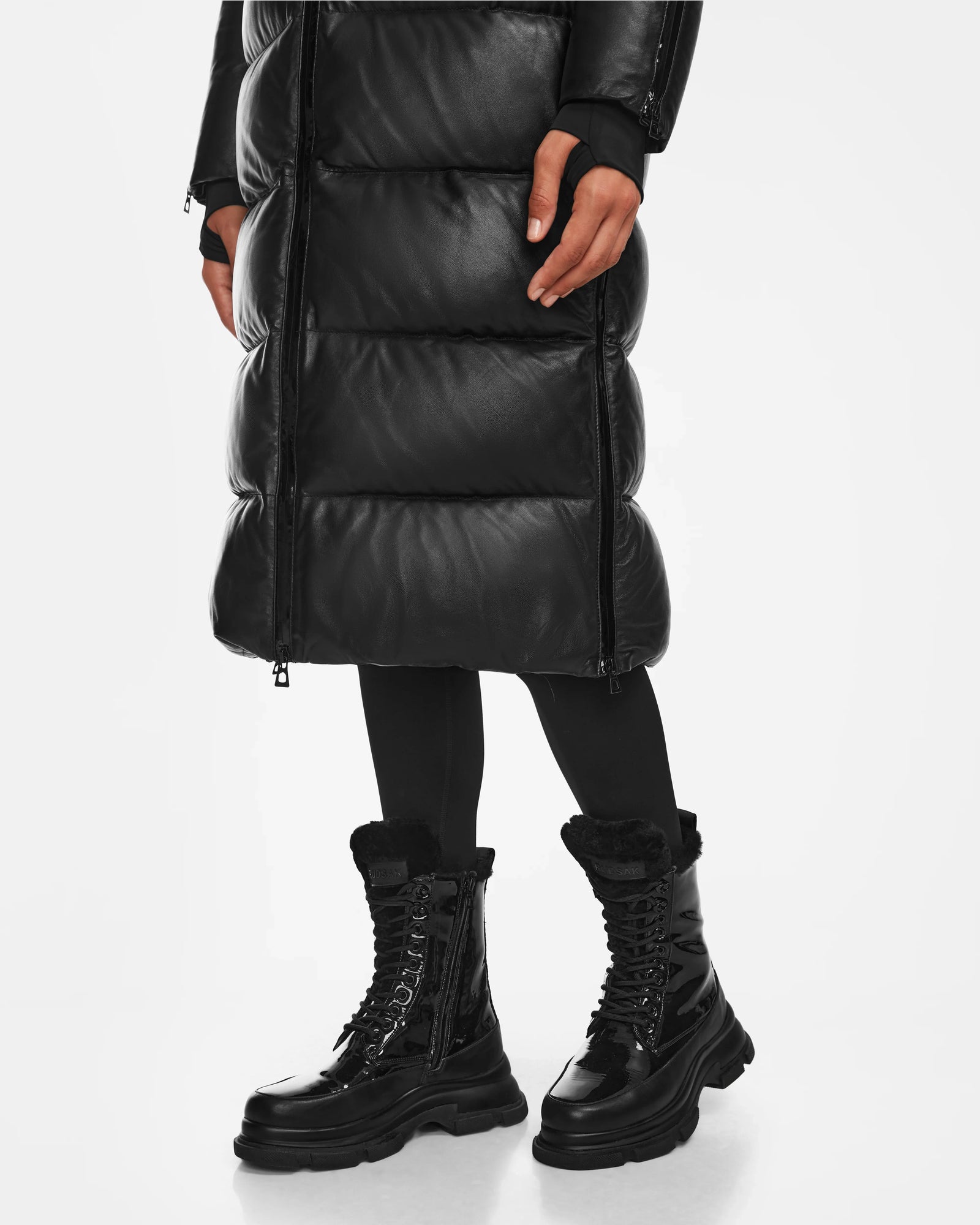 A person in a long black puffer coat, leggings, and Rudsak’s LANA - 8223506 BLACK mid-calf lace-up boots with thick soles stands against a white backdrop, showcasing cozy women’s winter boots ideal for cold days.