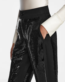 Someone wearing the Rudsak LEAR - 8524553 Black sequin pants with a long-sleeve white top, hand in pocket, shown waist-down against a plain background.